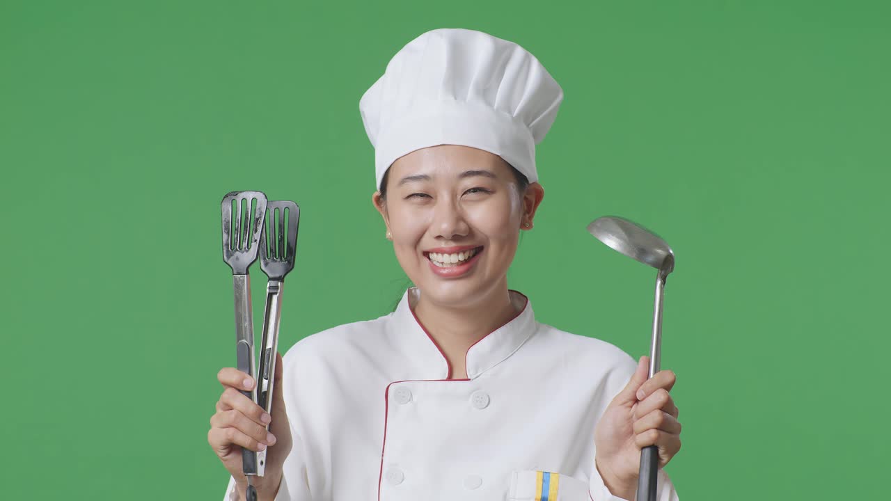Close Up Of Asian Woman Chef Smiling And Showing Tongs And Ladle To Camera While Standing In The Green Screen Background Studio