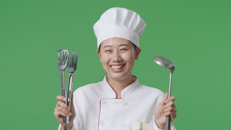 Close Up Of Asian Woman Chef Smiling And Showing Tongs And Ladle To Camera While Standing In The Green Screen Background Studio