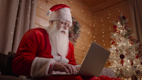 the real santa claus bottom view sits with a laptop on a couch with glasses and works at the laptop. christmas and christmas eve sonta klaus works at home with garlands and a laptop in a decorated