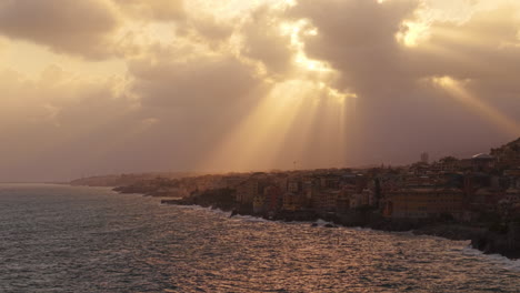 Coastal Mediterranean city near sea drone aerial view with golden sun rays breaking through thick clouds in sky during sunset