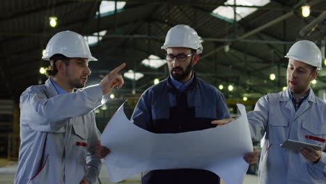 three engineers wearing helmets holding blueprint and tablet while talking and walking in a factory