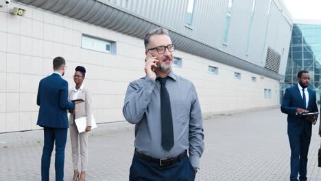 gray-haired businessman wearing glasses and talking on the phone while walking down in the street