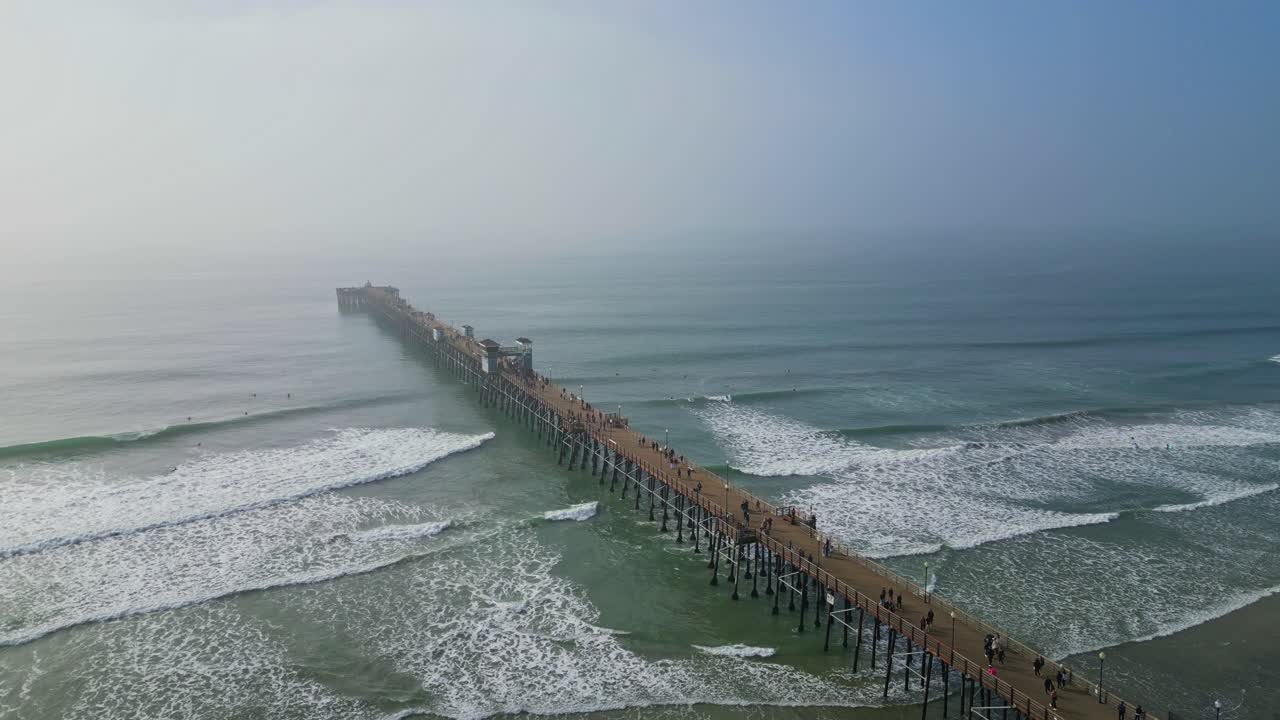 A drone shot of a California coastal pier during the day, featuring crashing waves, surfers, and crowds walking along the pier.