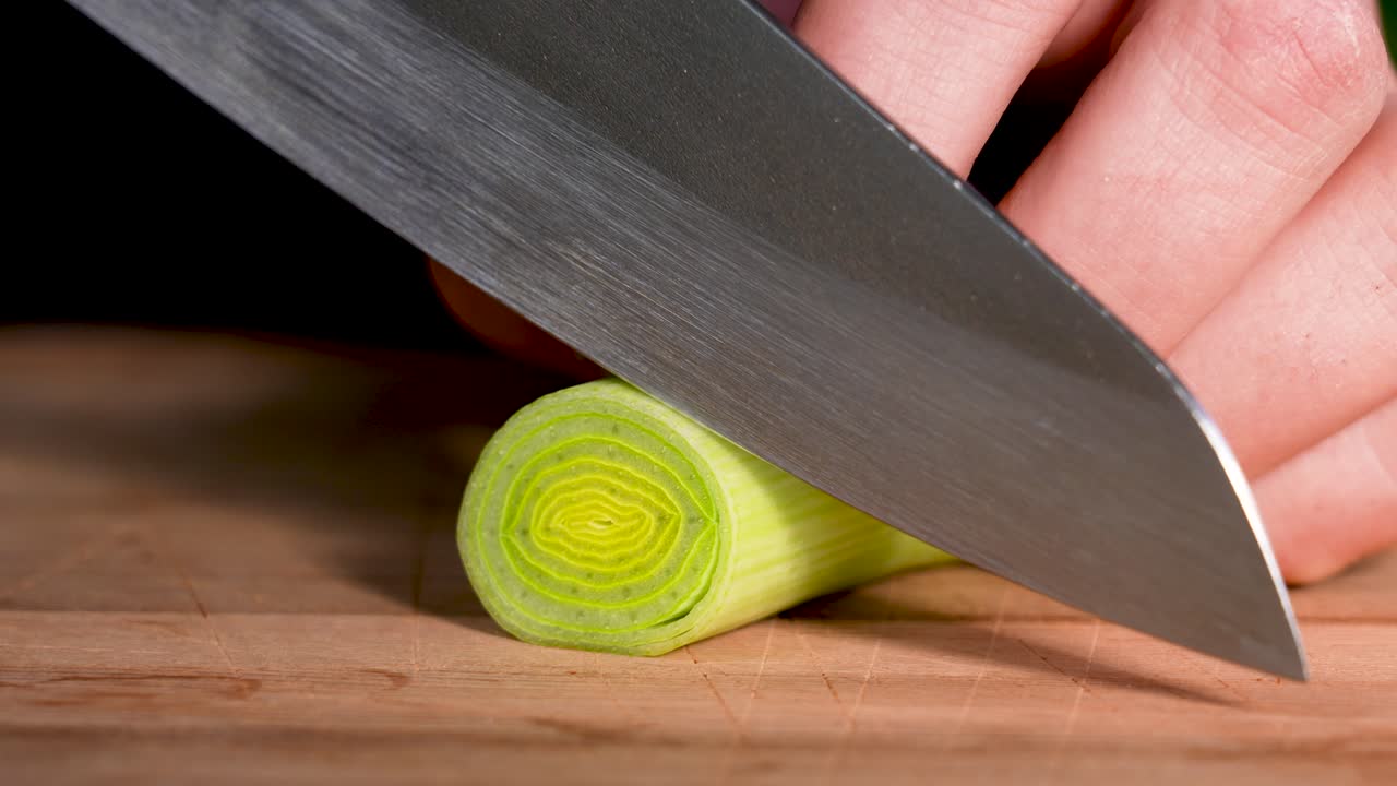 A macro close-up shows a knife smoothly slicing through a fresh leek on a wooden cutting board. Detailed layers and precise motion highlight clean food preparation and natural kitchen aesthetics