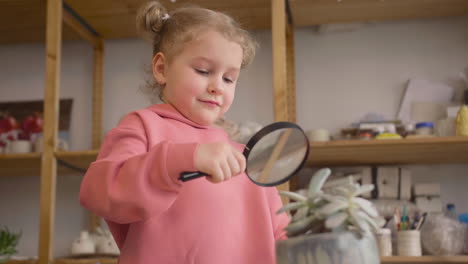 Vista-Inferior-De-Una-Niña-Observando-Una-Planta-Con-Una-Lupa-Sentada-A-La-Mesa-En-Un-Taller-De-Artesanía