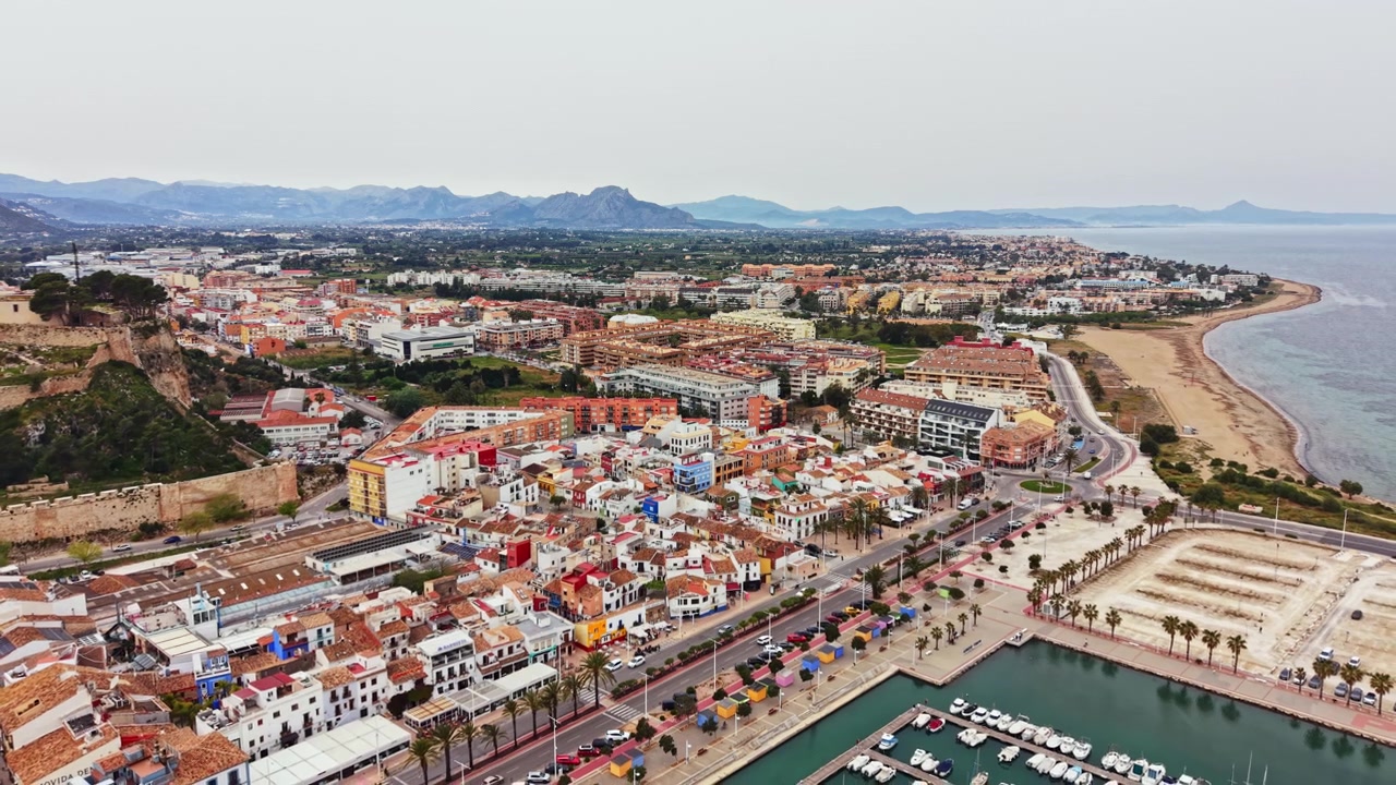Luftfoto af en kystby i Spanien, der viser bygninger, gader og landskab langs kystlinjen i dagslys
