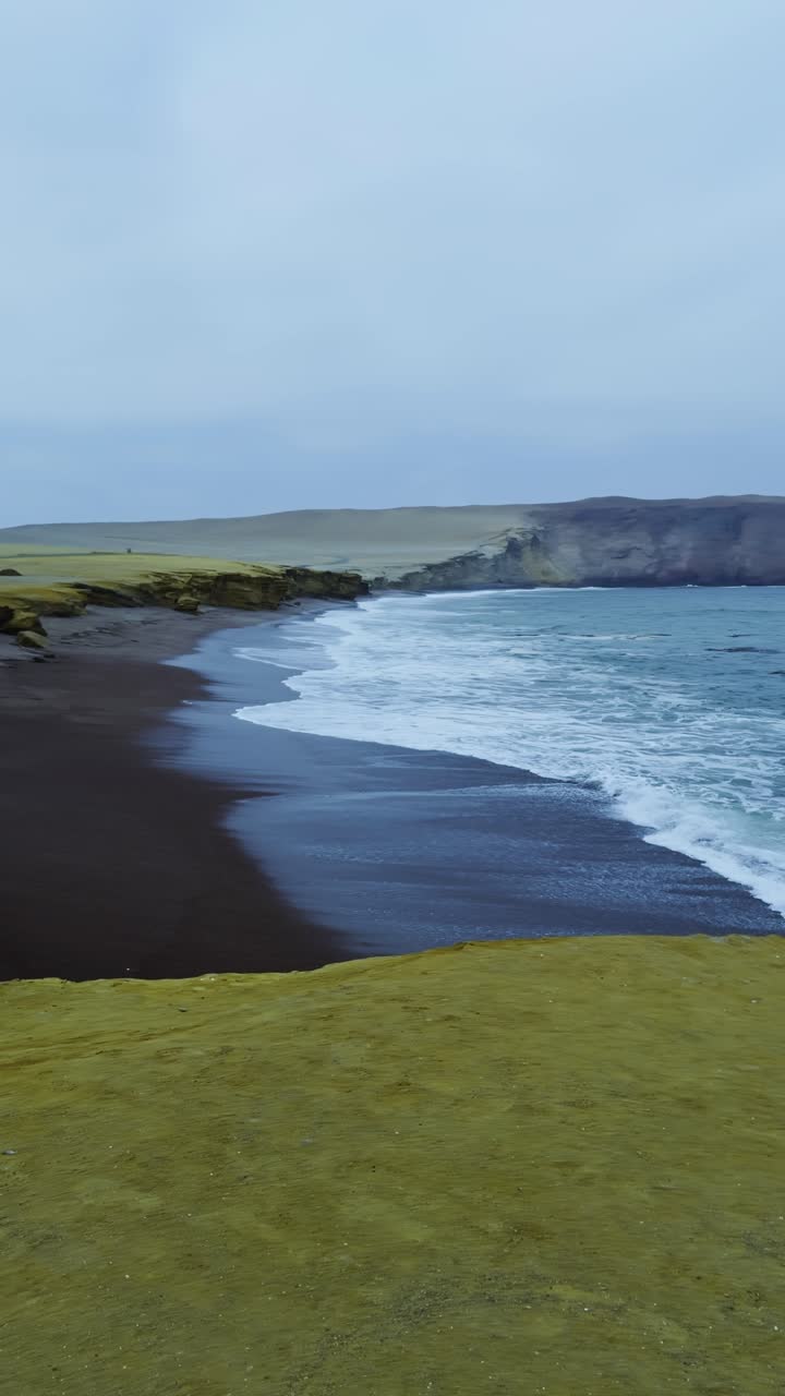 Sweeping wave breaks and recedes across Playa de Rojas coastline with ripples and mist on black sandy shore and cliffs, vertical establishing backdrop