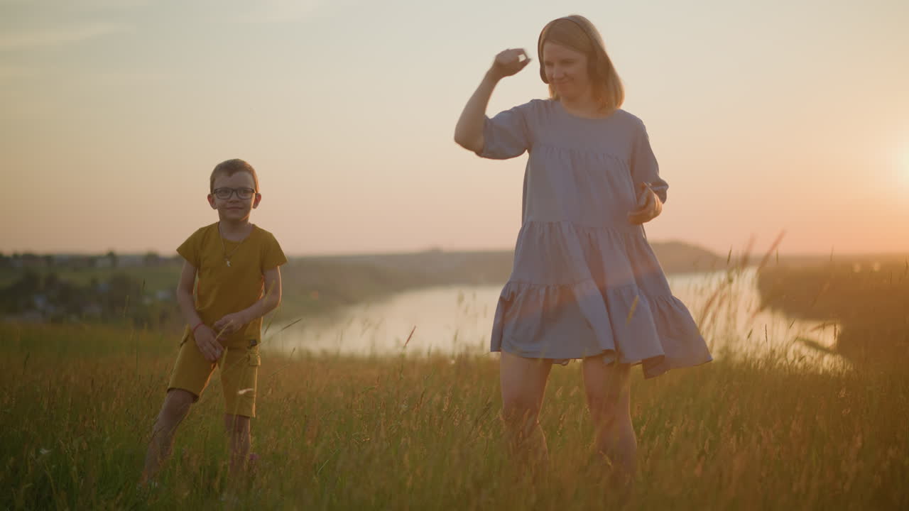 파란색 드레스를 입고 헤드을 착용 한 여성은 황색 옷을 입은 아들과 함께 춤을 추고, 해가 지는 동안 평화로운 호수 의 경치 좋은 풀에서 기과 연결의 순간을 포착합니다.