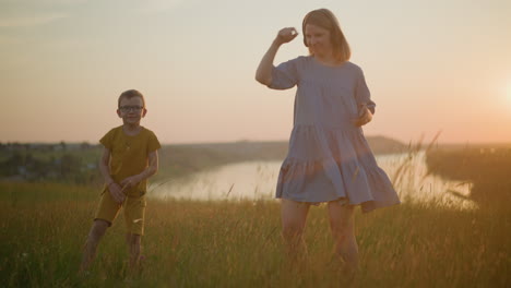 una mujer con un vestido azul, con auriculares, baila con su hijo vestido de amarillo en un pintoresco campo de hierba al lado de un lago sereno al atardecer, capturando un momento de alegría y conexión