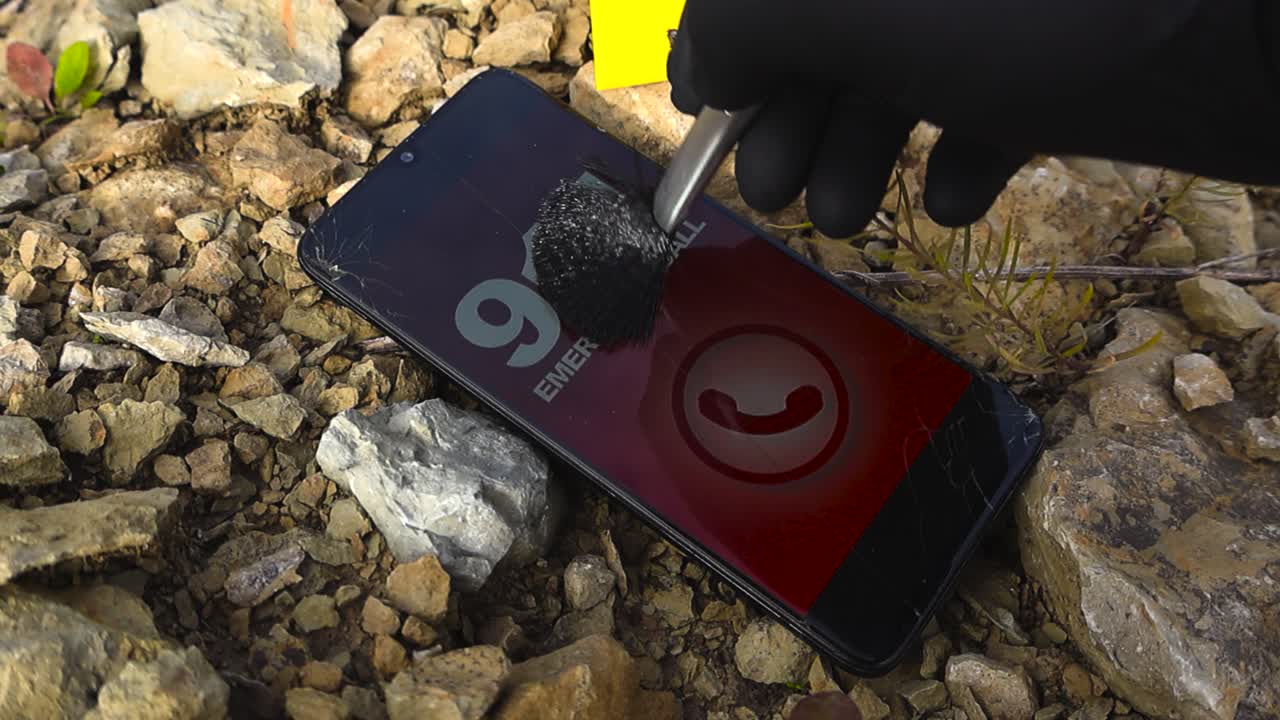 Close up view of a police detective or investigator dusting for fingerprints on a cell phone that is calling 911 emergency number on gravel ground next to police yellow forensic crime marker