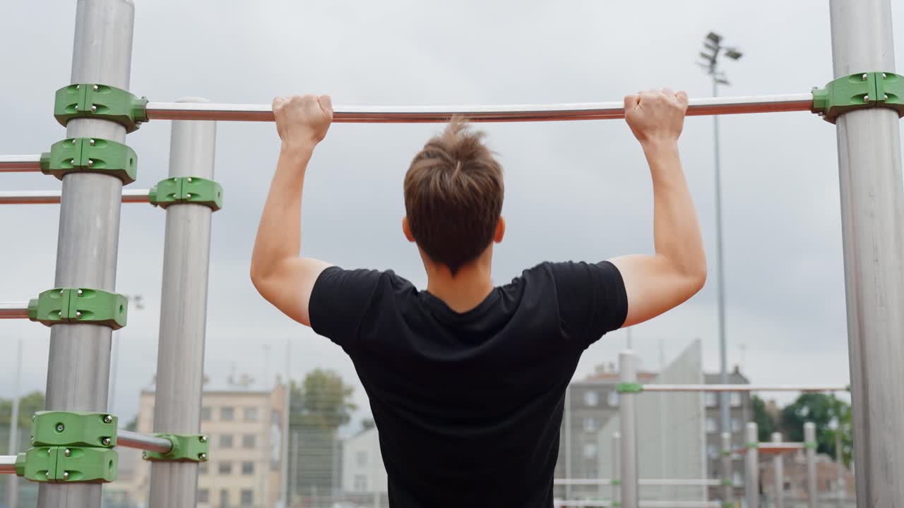 Young man performing a pull up exercise on a horizontal bar at an outdoor gym, building strength and working out for fitness and physical health, rear view, static camera, slow motion