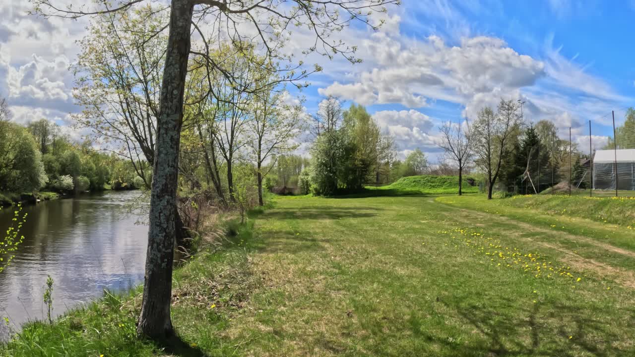 First person point of view walk through a path winds past lush green grass (Poaceae) and diverse trees under a bright sky. This immersive footage conveys the experience of exploring scenic urban park