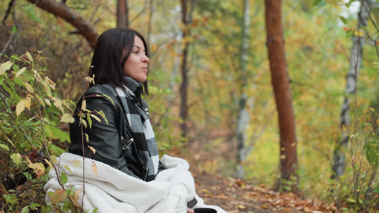 white woman sitting in autumn forest wrapped in blanket and wearing leather jacket and scarf, gazing into distance with reflective expression, leaves falling around, birch and pine trees