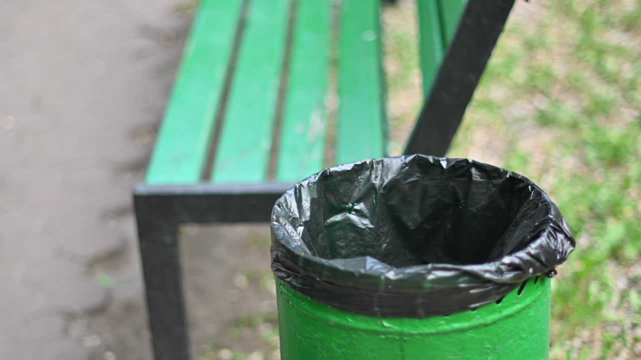 Woman throwing out a plastic cup in a green trashcan, near a bench in the park