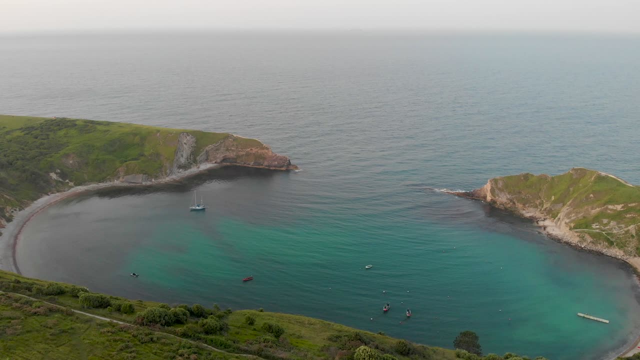 una imagen aérea de la bahía de lulworth con playa de arena, agua azul cristalino, barco de pesca, kayakistas y olas que se estrellan en el acantilado rocoso
