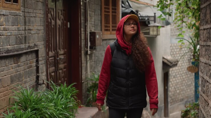 Donna asiatica che cammina in uno stretto vicolo del villaggio, viaggiatrice incappucciata si ferma a studiare edifici in pietra e porte in legno, vegetazione che si arrampica sui muri, indossa felpa rossa, gilet nero, berretto giallo e occhiali, curiosa.