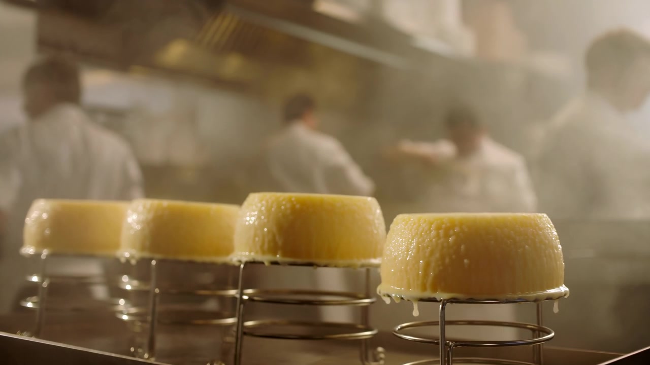 Row of small round custard desserts inverted on chrome ring holders in foreground, glossy tops and dripping edges showing cooling and setting after heat removal, with steam, blurred kitchen staff