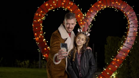 father and daughter taking a selfie in front of a heart-shaped light display