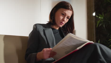 businesswoman wearing gray suit reading documents in red folder with serious thoughtful expression while sitting on office couch under daylight, reviewing paperwork or analyzing business information
