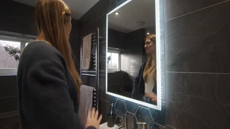 Modelo-Femenino-Mirando-En-Un-Espejo-Retroiluminado-Del-Baño-Ordenando-Su-Cabello