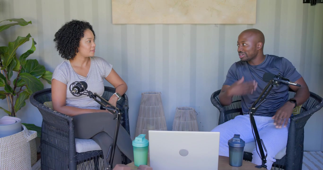 Anfitriones afroamericanos grabando en el porche, el hombre iniciando la discusión y gesticulando, la mujer moviendo los micrófonos.