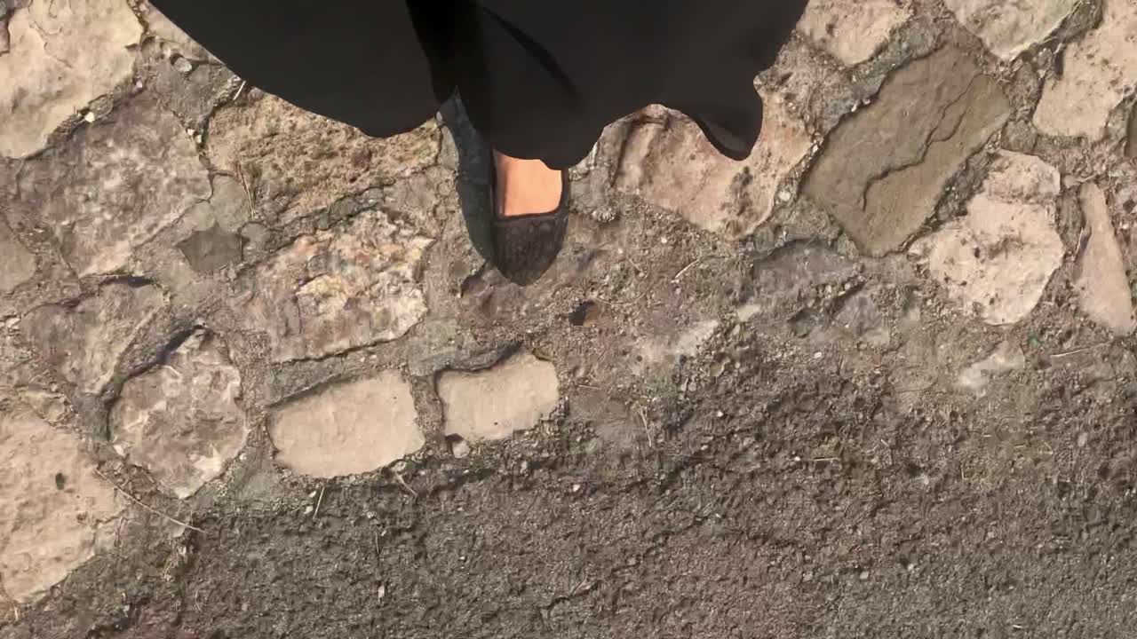 Faceless self-portrait  of woman walking on paved cobblestones on breezy day in black silk dress in 4k