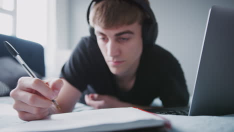 male college student wearing headphones works on bed in shared house with laptop