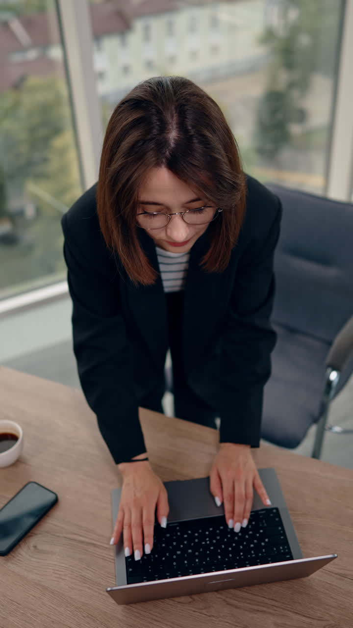 Brunette lady types on the laptop keyboard standing at the desk. Woman rises head and shows thumb up to camera. Top view on the female employee working in the office. Vertical video.