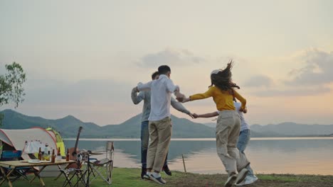 group four people friends asian men and women camping picnic celebrating holding hands in a circle and dancing having fun and enjoying ground tent. reservoir area during sunset vacation time.