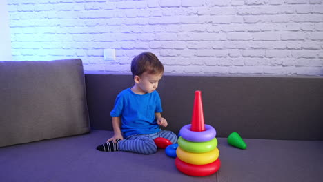 calm little baby boy wearing blue t-shirt playing with toy sitting on the sofa. cute toddler assembling the pyramid.