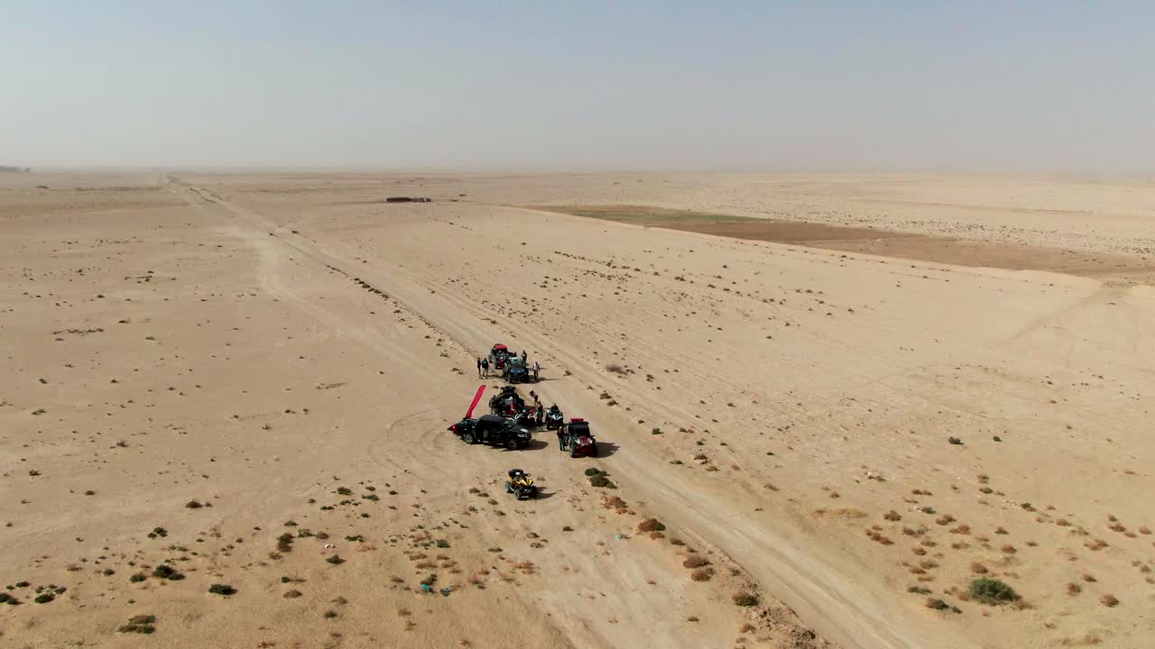 Ripresa aerea con drone di un gruppo di quad e buggy fuoristrada fermi lungo un sentiero sabbioso nel deserto in Tunisia, con montagne velate e un vasto paesaggio arido sullo sfondo.