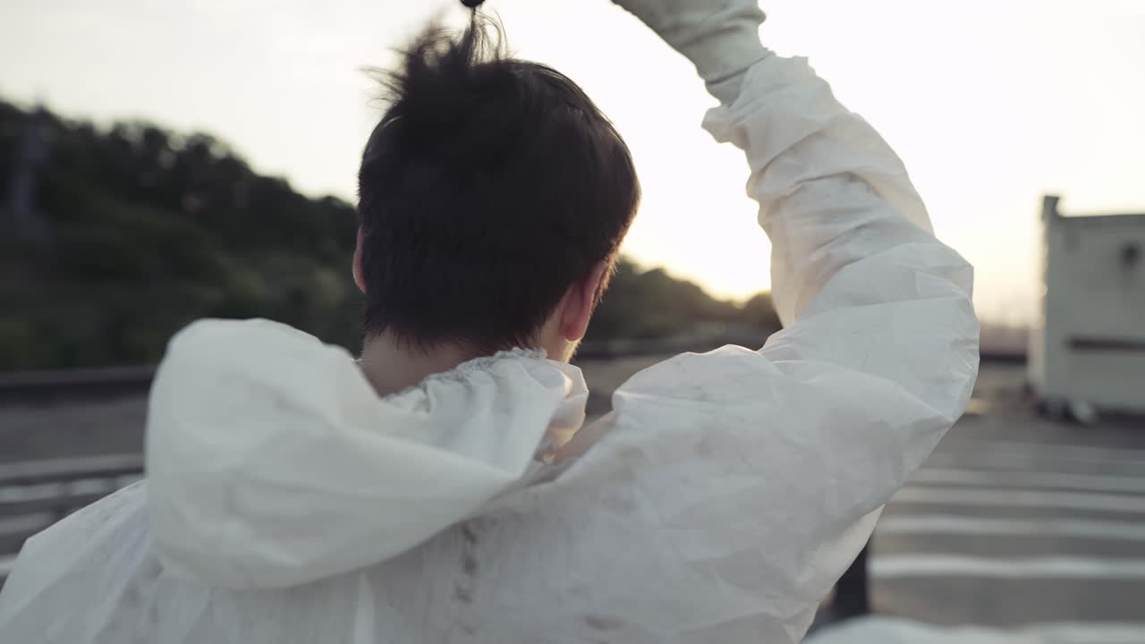 지붕 위에 서 있는 보호복을 입은 남자가 호흡기와 마스크를 벗고 뒤로 보입니다.