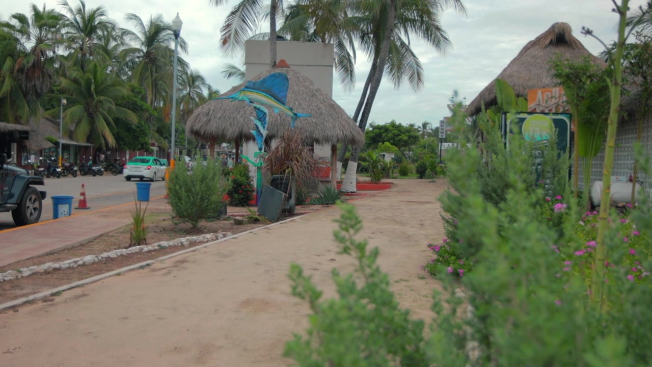 Una rilassata città surfista hippie a Oaxaca, Messico, con palme, sabbia, capanne di bambù e ristoranti, in una giornata nuvolosa e coperta, al rallentatore.