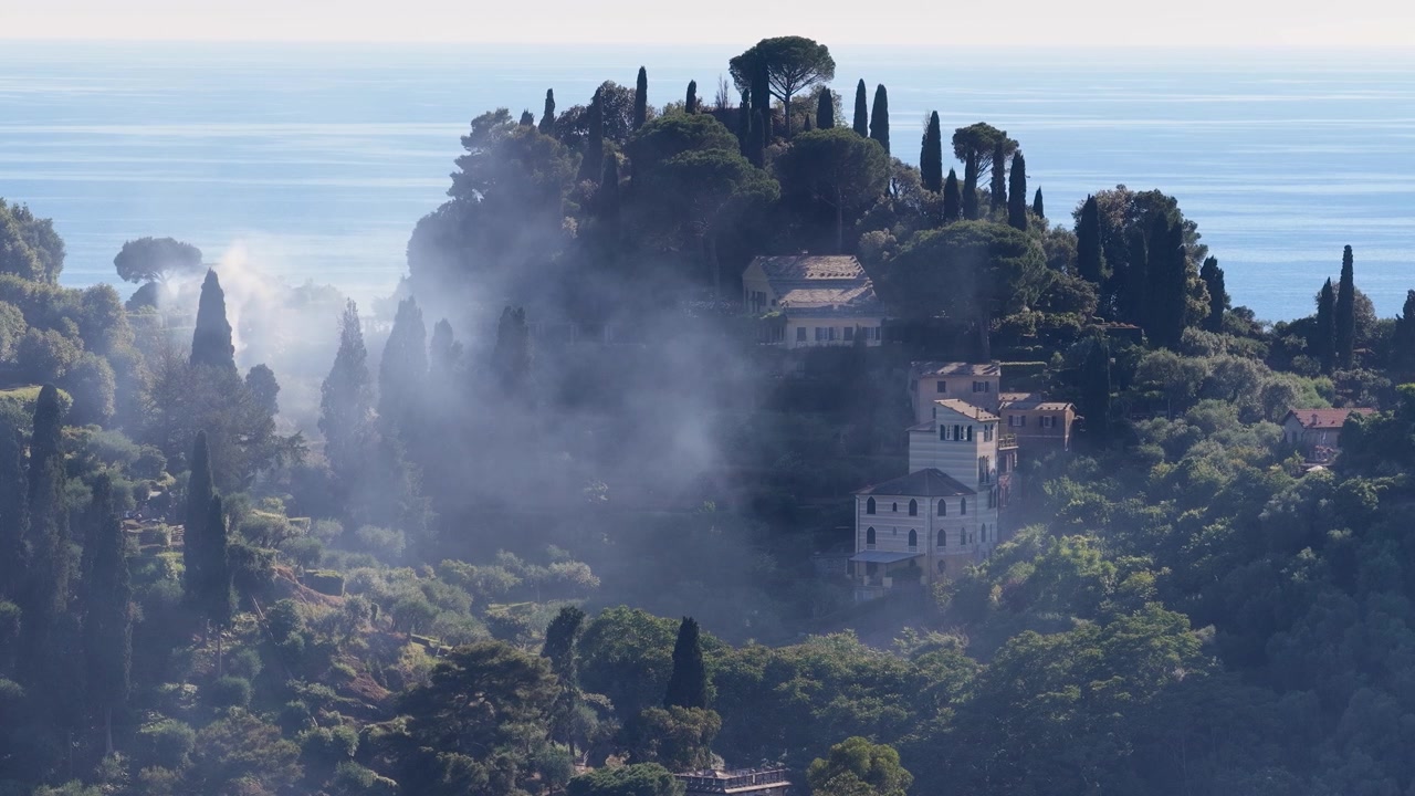 La nebbia avvolge un lussureggiante pendio verde con ville tradizionali che si affacciano sul mare azzurro.