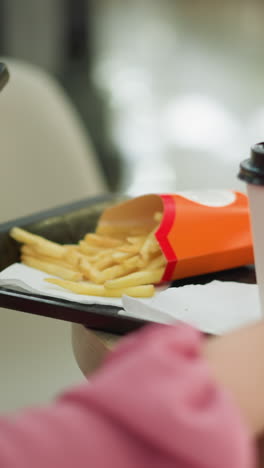 una mujer ajusta una bandeja negra en una mesa de madera que contiene una taza de café, una hamburguesa y patatas fritas con la mano izquierda mientras sostiene su teléfono en la mano derecha, el fondo está suavemente borroso