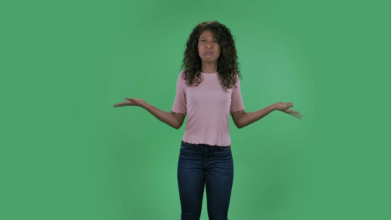 Porträt einer schönen jungen afroamerikanischen Frau, die genervt gestikuliert und Irritation und Wut ausdrückt. Eine Brünette mit welligem Haar in Jeans und einer beigen Bluse vor einem Greenscreen im Studio.