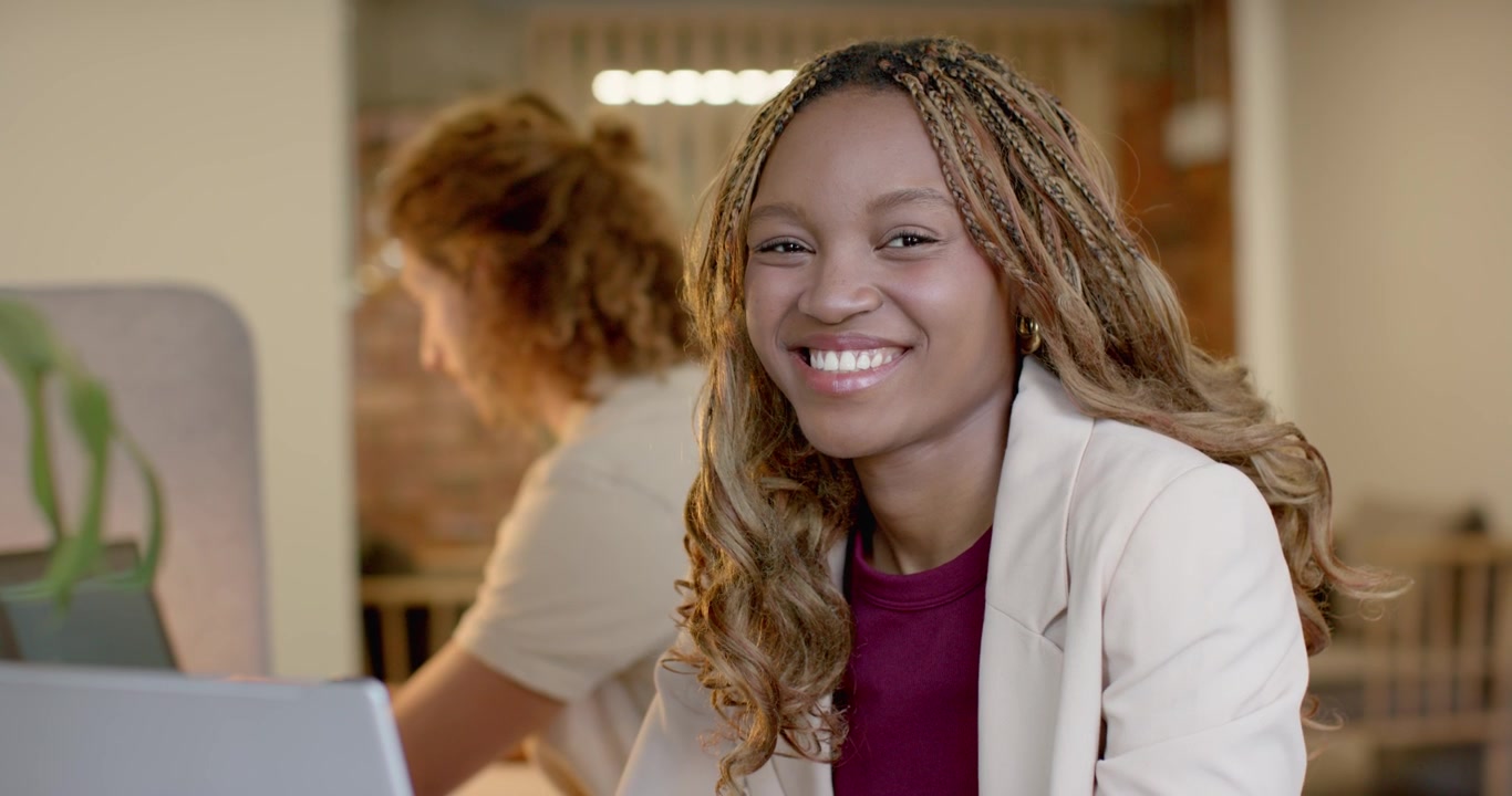 Donna afroamericana che alza lo sguardo con la telecamera che si avvicina, sorridendo e chinandosi verso il laptop sulla scrivania