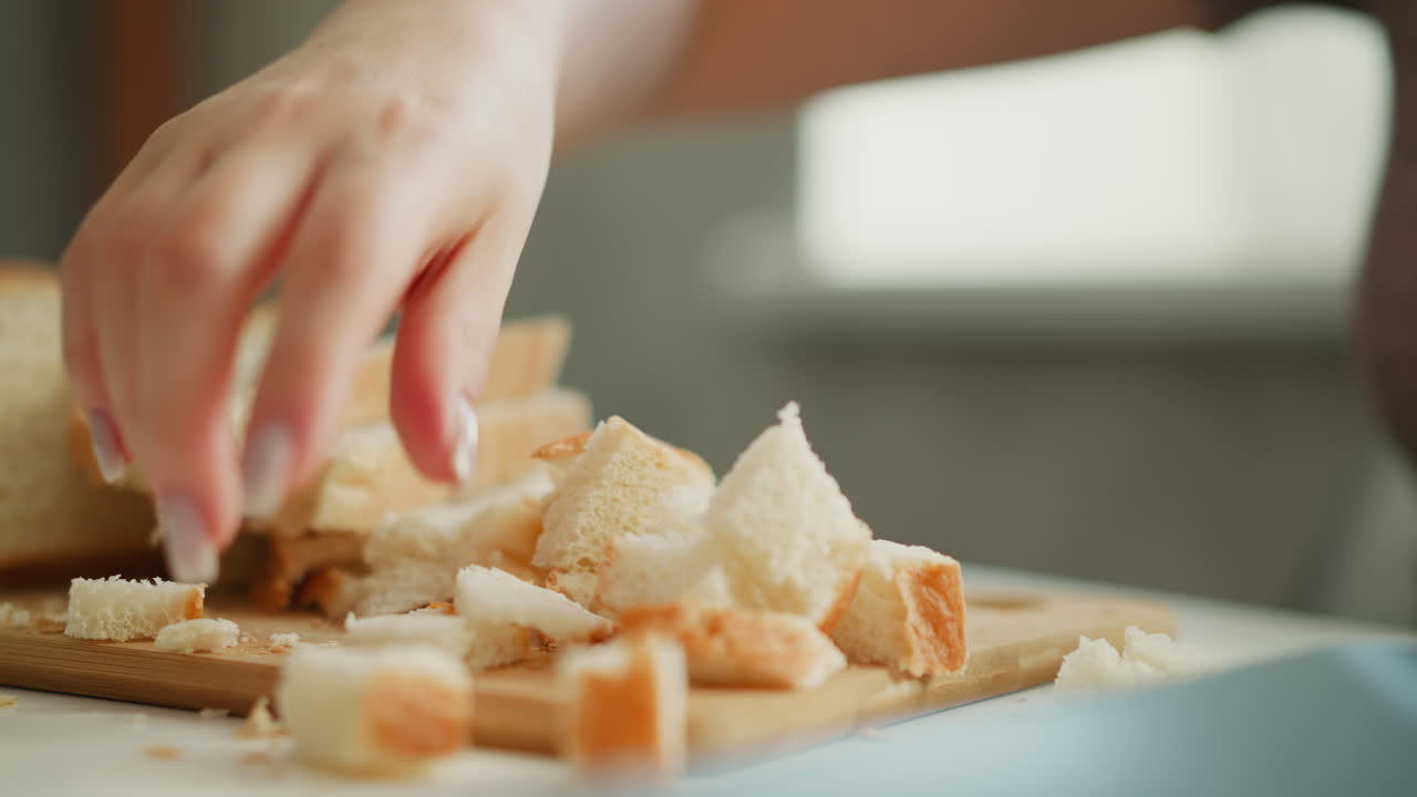 Female chef lifting pieces of bread loaf with delicate hand gesture on wooden cutting board, crumbs falling onto white table under soft morning window light, close up shot capturing texture