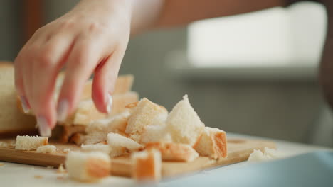 female chef lifting pieces of bread loaf with delicate hand gesture on wooden cutting board, crumbs falling onto white table under soft morning window light, close up shot capturing texture