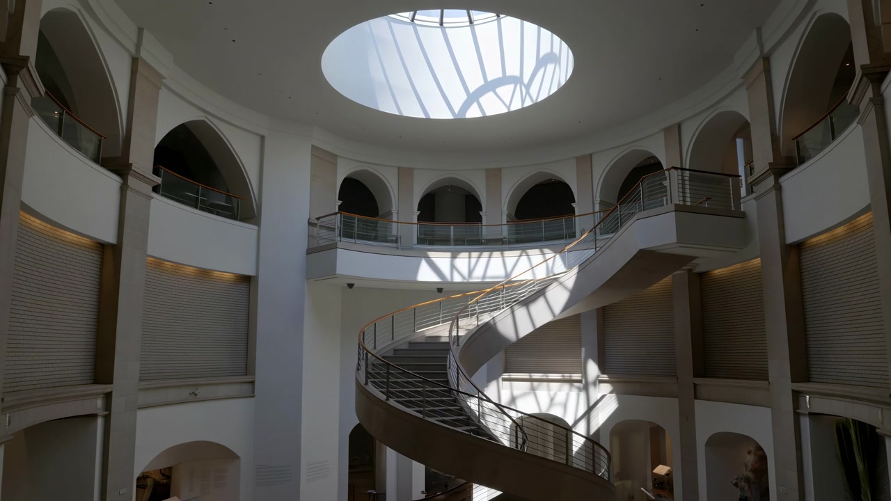 Central spiral ramp and sweeping curved balcony fill multi-level atrium beneath circular skylight, while directional sunlight creates shifting linear shadows across stair surfaces and bright