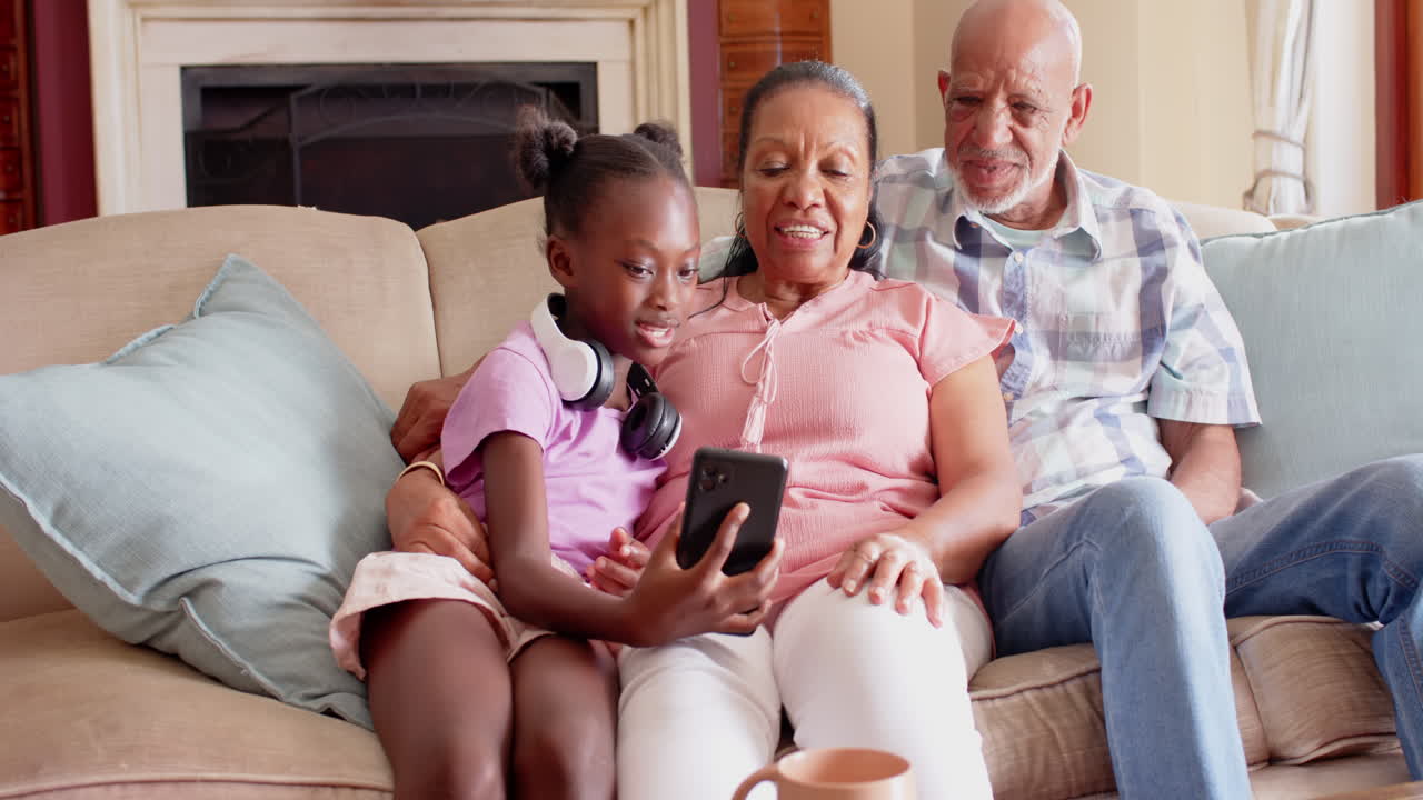 Laughing together, diverse grandparents and child looking at smartphone on couch at home
