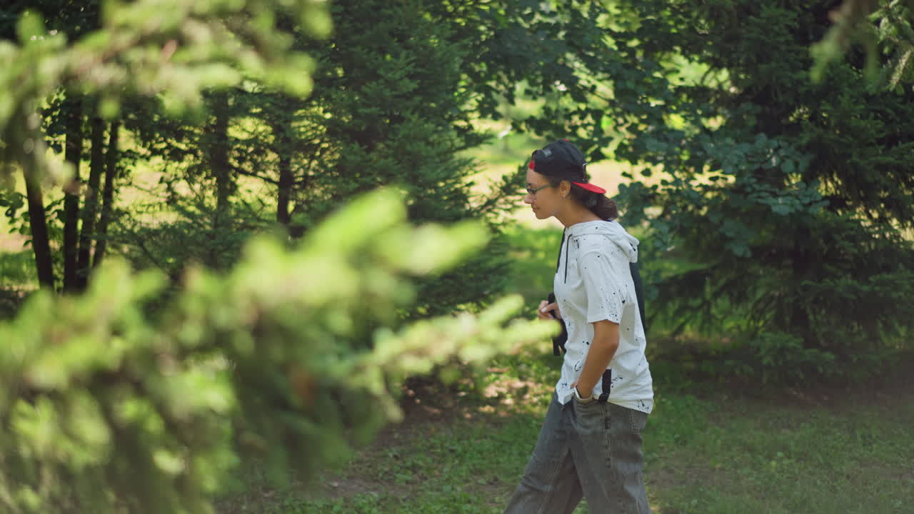 Latina explorer pausing beneath green canopy, handsonhips survey of parkland, confident stance with shoulder bag, dappled light through branches, casual summer outfit, observational mood and candid