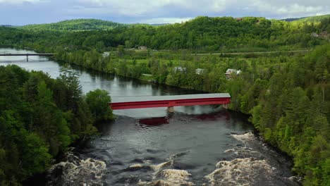 Drone-shot-of-a-picturesque-red-covered-bridge-over-the-Gatineau-River-in-a-tree-covered-valley-of-Wakefield-Quebec-Canada