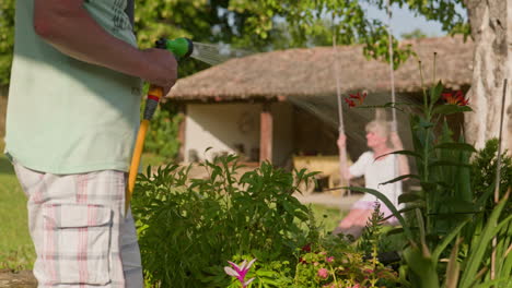 Gardner-waters-plants-on-summer-morning-as-woman-relaxes-on-garden-swing