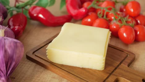 a block of cheese on a cutting board with tomatoes, red peppers, and garlic.