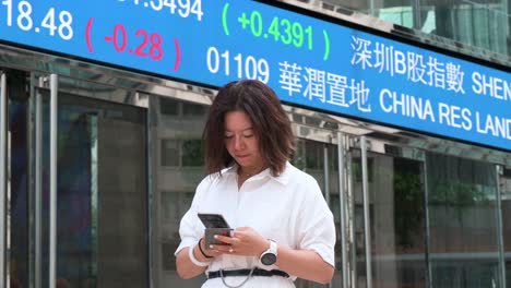 from a low-angle perspective, a female executive is seen using her smartphone in exchange square (hkex), the home of the hong kong stock exchange, while company stock ticker symbols flash behind her.
