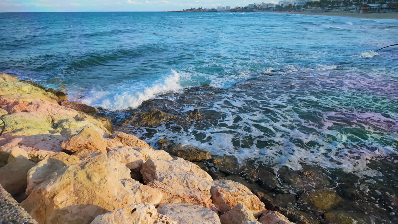 Onde che si infrangono contro una costa rocciosa con massi dorati sotto un cielo sereno, probabilmente in una regione costiera di Cipro.