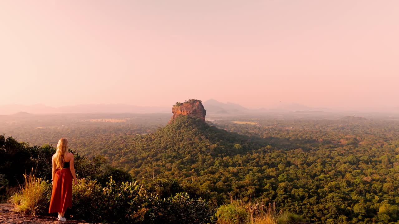A serene and cinematic moment as a woman sits atop Pidurangala Rock, gazing at the majestic Sigiriya Rock illuminated by the first light of sunrise.