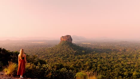 a serene and cinematic moment as a woman sits atop pidurangala rock, gazing at the majestic sigiriya rock illuminated by the first light of sunrise.