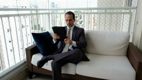 man in a suit and tie sitting on the sofa and making a video call with his tablet.